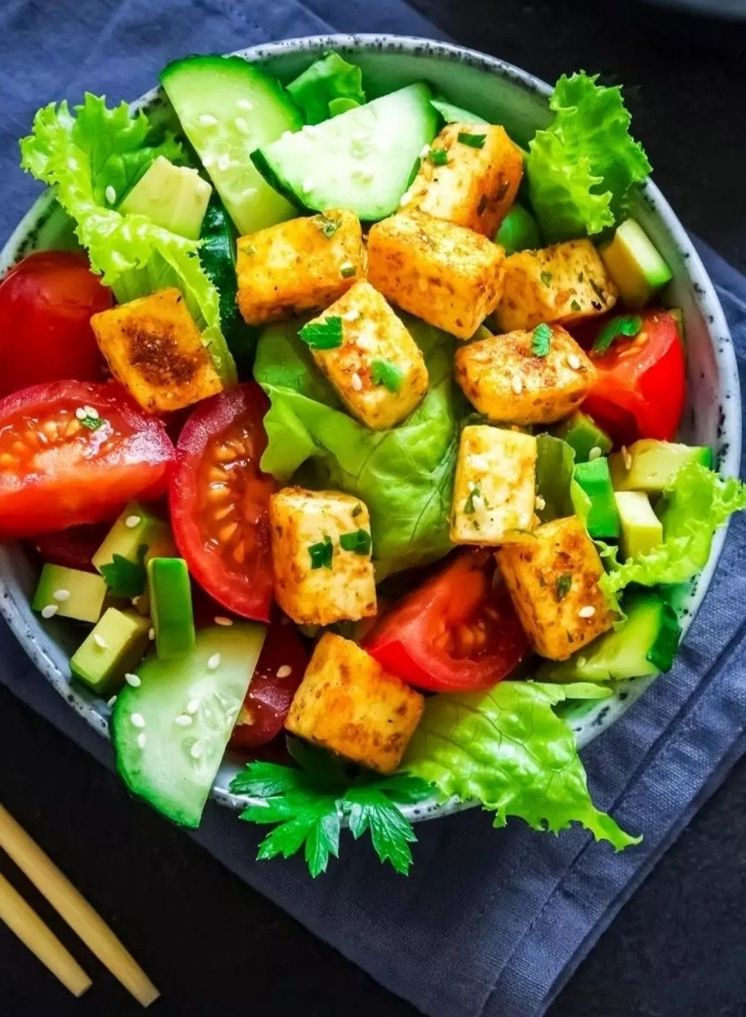 Fresh salad bowl with seared tofu, tomato, cucumber, avocado, and greens beside wooden chopsticks